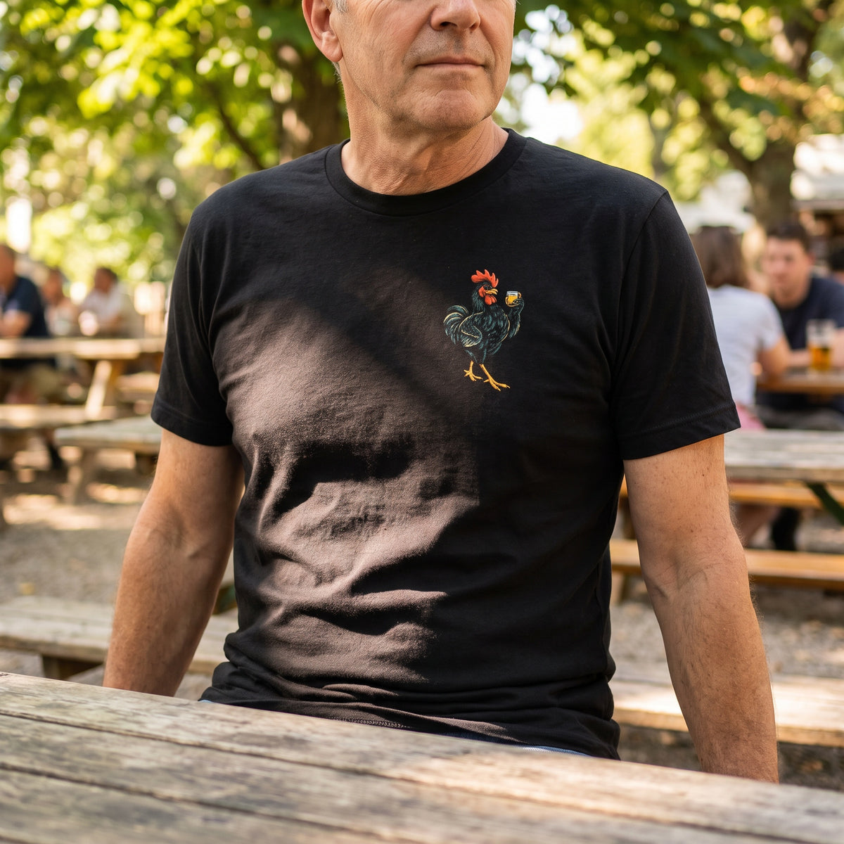 Man wearing the black Whisky Business rooster T-shirt with a small whisky-glass chest illustration at an outdoor table.
