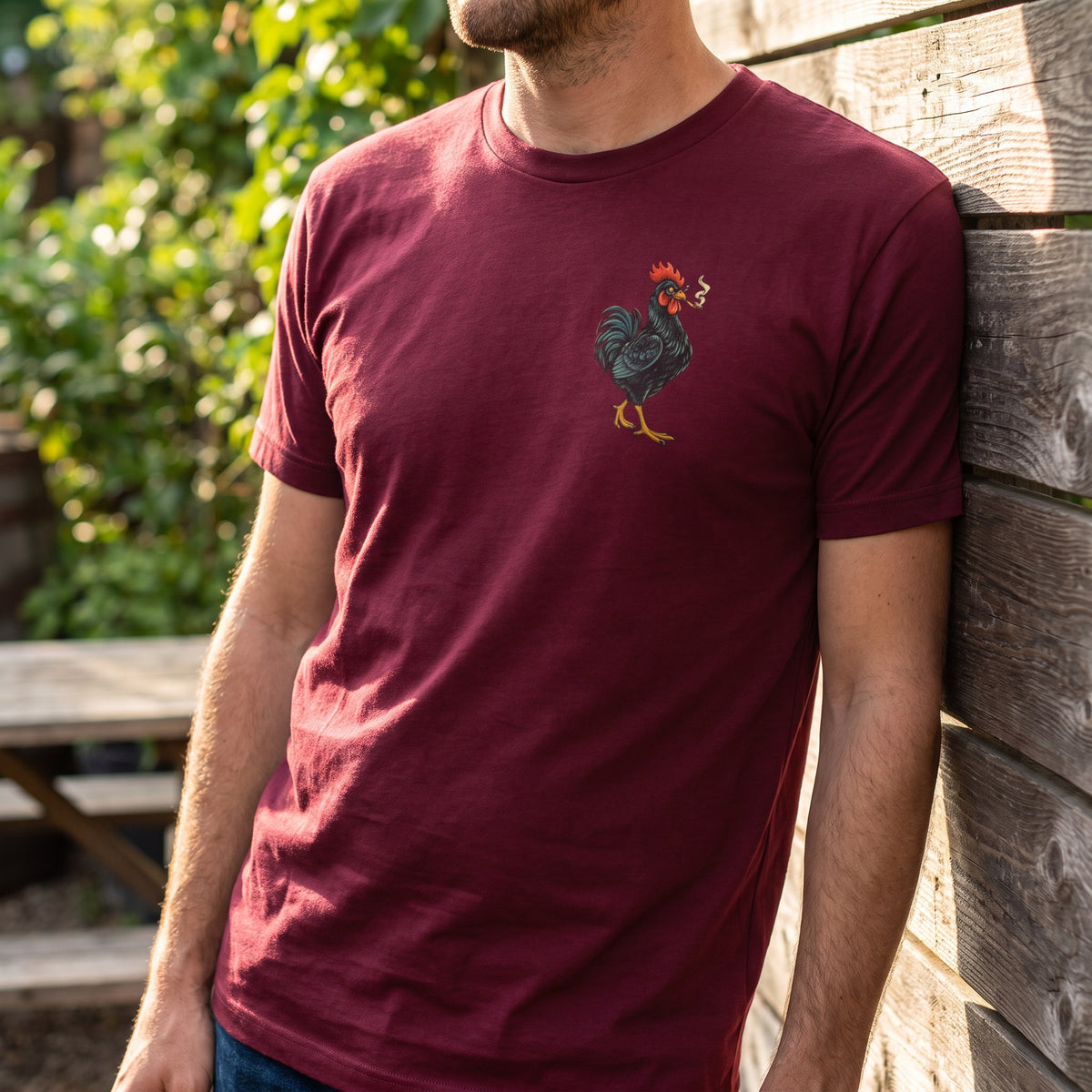 Man wearing the cardinal red You Can Call Jay T-shirt with a small joint-smoking rooster chest graphic beside a wooden wall outdoors.