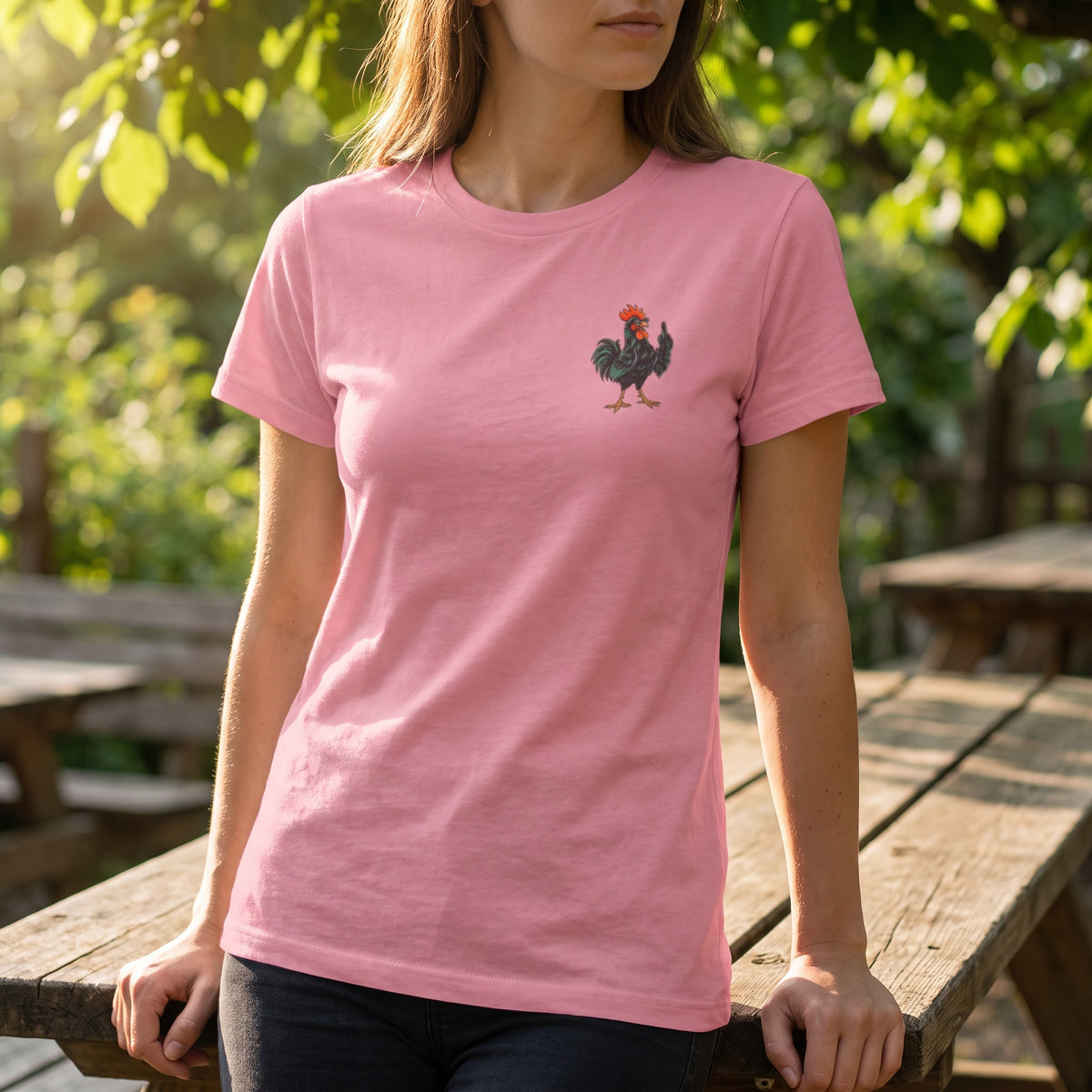 Woman wearing the light pink Giving the Bird T-shirt with a small middle-finger rooster chest graphic beside an outdoor wooden railing.