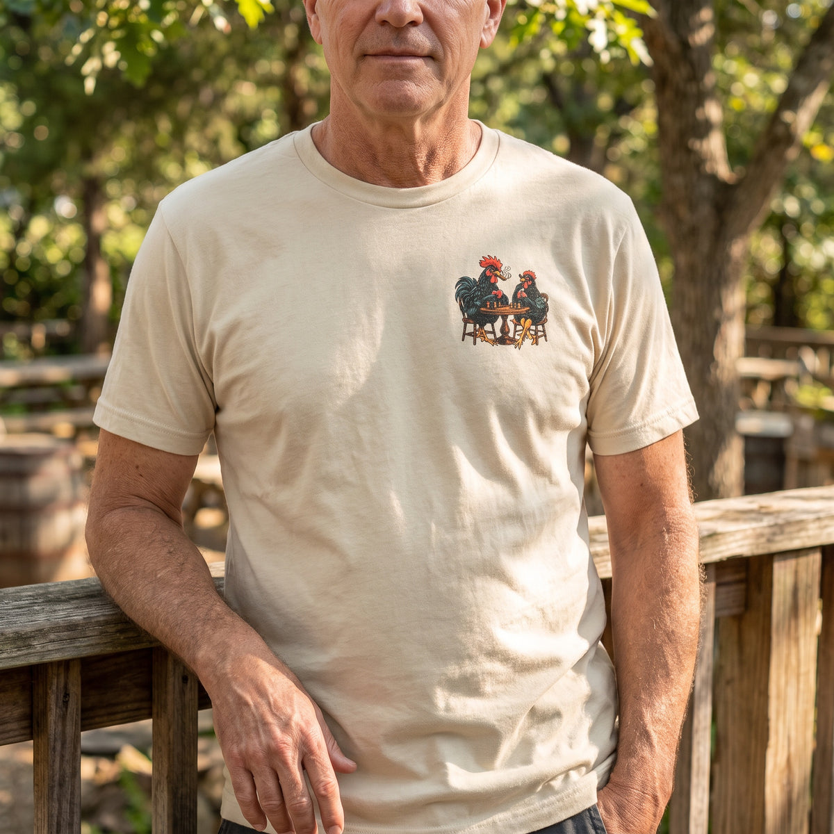 Man wearing the cream High Stakes, Low Clucks T-shirt with a rooster-and-hen card game chest graphic beside a wooden railing outdoors.